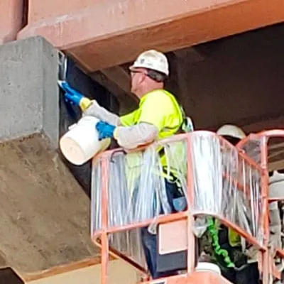 Technicians in a lift applying a carbon fiber composite wrap to portions of the supports below a bridge