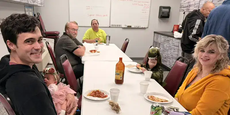 Several people having lunch in the company break room and a couple of them are dressed up for halloween