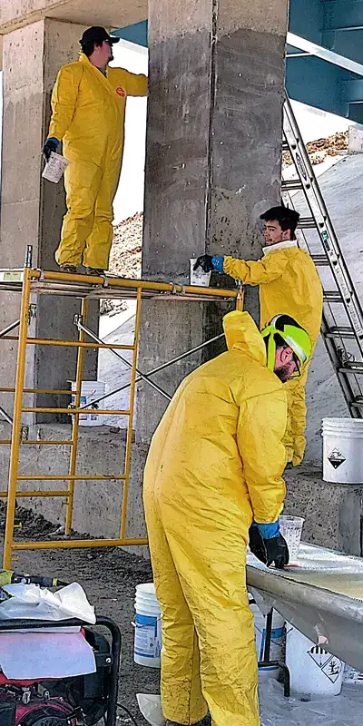 Several technicians wearing tyvek and using scaffolding to apply fiber wrap to a bridge column