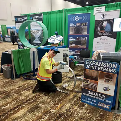 Caleb kneeling on the ground at a tradeshow preparing to install HydraTite internal pipe joint seal