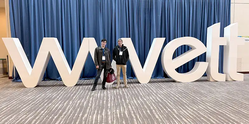 Two people sitting in front of a large WWETT sign