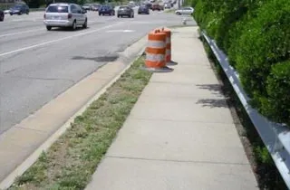 A walkway near a road with traffic cones marking where a person could enter the pipe below