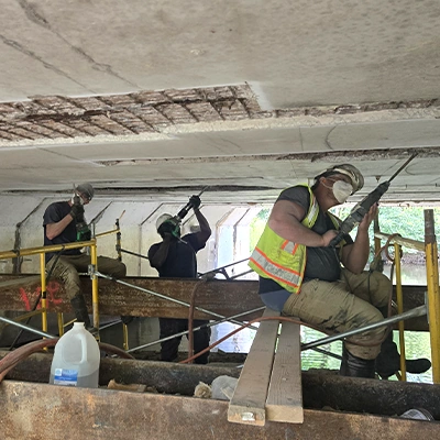 Several field services technicians working above a creek to remove loose concrete from the underside of a bridge so that it can be patched