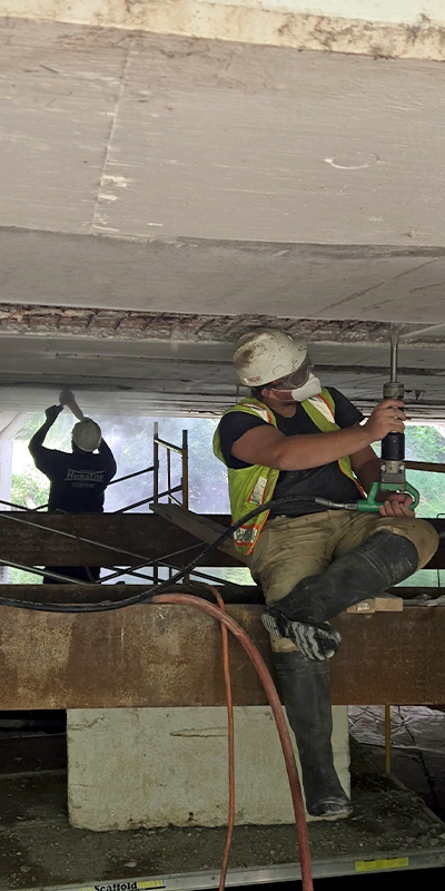 Two technicians working under a bridge to chip out loose concrete